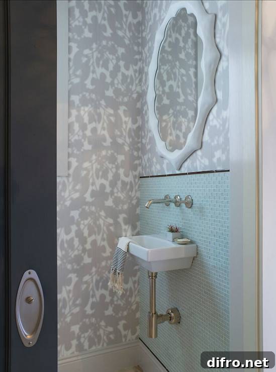 Chic powder room features a black door opening to reveal subtle white and gray floral motif wallpaper highlighting a modern white mirror mounted above an aqua blue glass mini brick tiled backsplash trimmed with black pencil tile framing a mini wall mount sink with modern wall mount faucet.