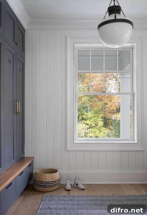 A black and white lantern lights a mudroom boasting a white vertical shiplap wall contrasting dark gray mudroom lockers adorned with brass hardware and fixed beneath dark gray overhead cabinets and over dark gray shoe drawers with a wooden bench top.