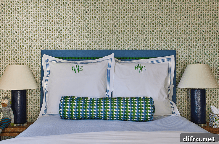 Green and blue monogrammed shams sit in front of a blue nailhead headboard and behind a blue and green bolster pillow placed on blue bedding.