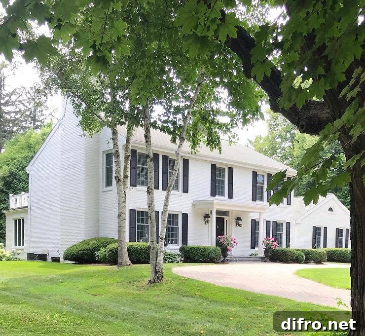 A circular driveway leads to a white brick colonial home contrasted with black shutters and accented with a portico finished with Greek columns lit by carriage lanterns.