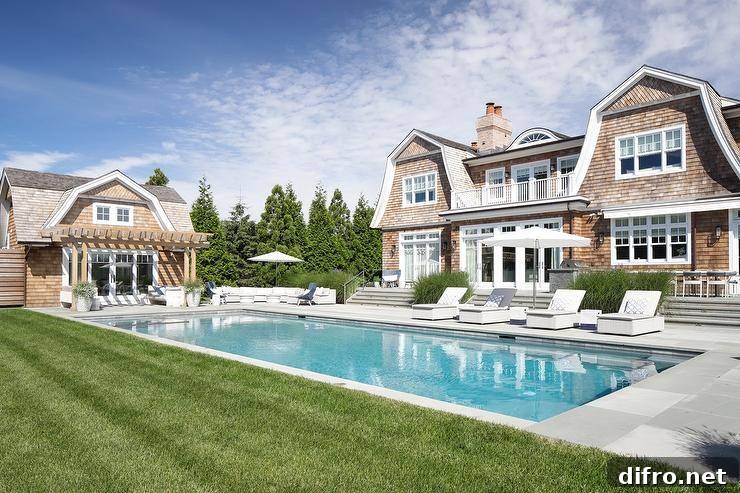 Cape Cod cottage with light brown shake siding and pool loungers by an in-ground pool.