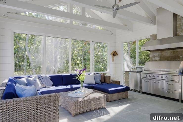 Beautiful sunroom with a white truss ceiling accented with a silver ceiling fan features a wicker sectional fitted with blue cushioned seats around a wicker ottoman as a coffee table atop gray pavers leading to the sunroom kitchen with a vent hood over plank backsplash and barbecue next to a stainless steel mini fridge.