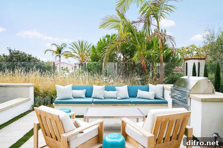 A built-in backyard bench topped with beautiful blue cushions accented with white pillows is positioned adjacent to a white outdoor kitchen finished with a stainless steel barbecue fitted to a concrete countertop. The sofa faces a stainless steel coffee table located in front of teak chairs topped with white cushions.