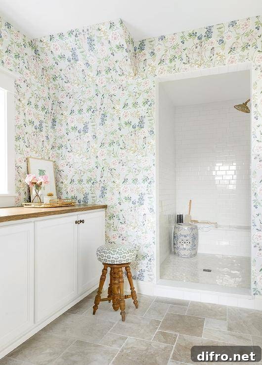 Clad in floral wallpaper, this welcoming bathroom features a doorless walk-in shower covered in white subway surround tiles and fitted with a subway tiled bench fixed behind a white and blue pierced drum stool. A wooden stool sits outside of the shower and in front of white cabinets topped with a brown wood countertop mounted beneath a window.