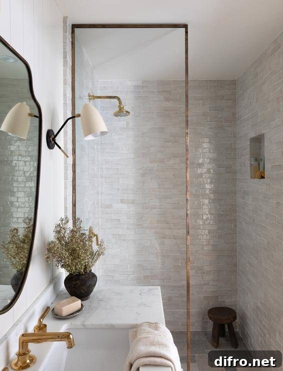 A glass partition with brass trim accents a shower clad in gray glazed surround tiles framing a niche located over a black stool. The shower is finished with a brass shower kit.