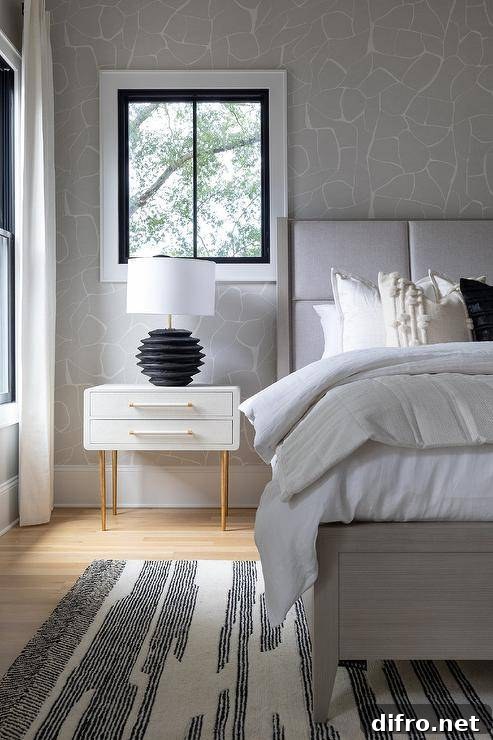 Bedroom features a light gray wood and fabric bed atop a black and white rug and a black lamp that illuminates a white and gold nightstand on white and gray wallpaper.