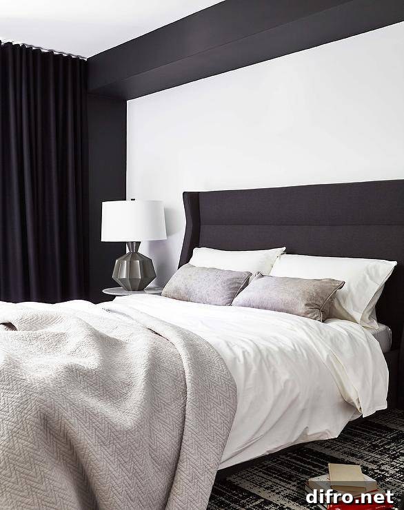A gray lamp lights a round marble top bedside table on a black and white rug beside a black wingback headboard.