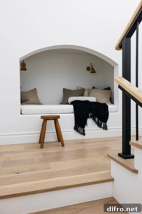 Brass sconces illuminated a staircase with built in reading nook accented with tan pillows and a black throw blanket and a wooden stool.