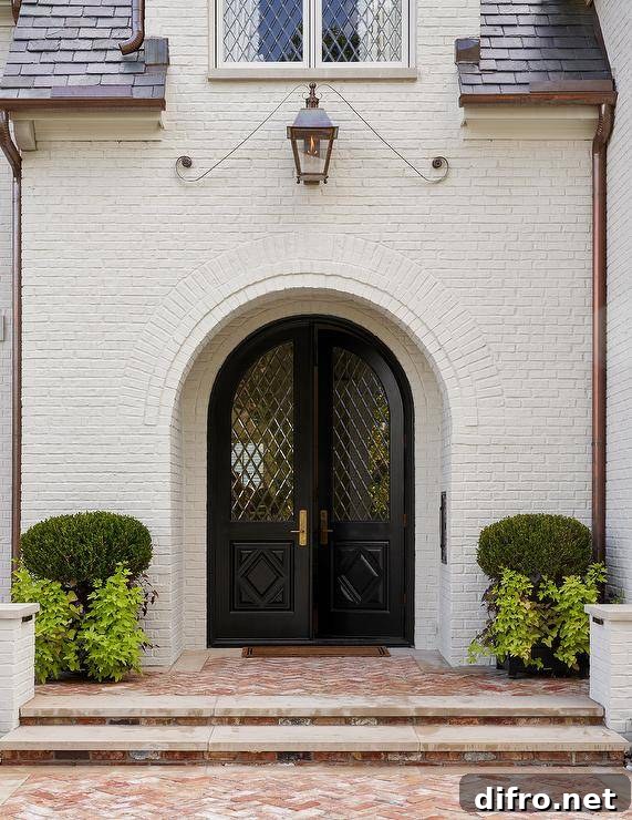 A white brick French chateau features black lattice front doors lit by a lantern and red brick herringbone pavers.