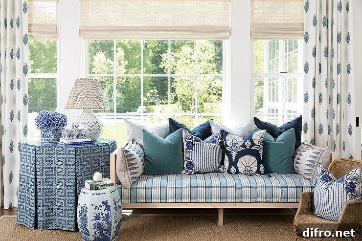 Paisley block print curtains hang from a window covered in bamboo roman shades hung above a cane day bed accented with a blue stripe cushion. The daybed sits on a bound jute rug beside an octagon skirted end table lit by a white and blue tiled chevron lamp.