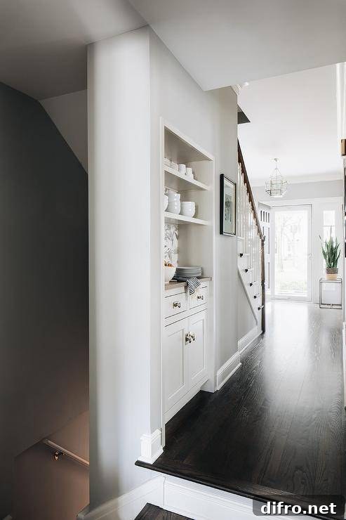 Built-in to the side of a staircase, a butler's pantry nook features white cabinets adorned with vintage hardware and fixed beneath stacked white shelves.