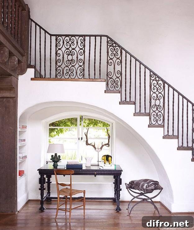 Mediterranean home features an arched alcove, placed under a wrought iron staircase, filled with a black desk and a wooden chair placed under arched casement windows situated next to built in shelving.
