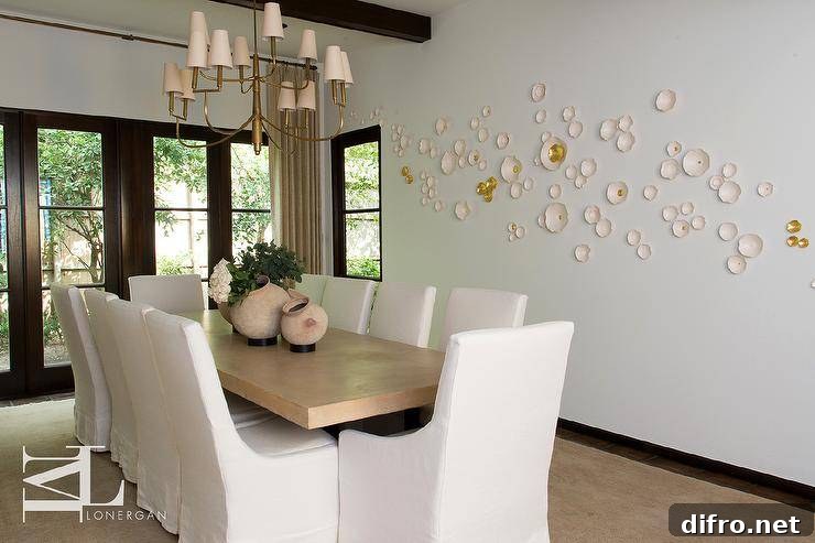 Elegant dining room boasts a gold dining table with white slipcovered dining chairs under an antique brass tiered Farlane Large Chandelier. White and gold wall decor on white walls is trimmed with black crown moldings under black wood ceiling beams in a lovely contemporary dining room design.