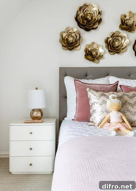 Pink and gray girl's room features a curved white nightstand accented with brass knobs and a hammered gold lamp and positioned beside a charcoal gray upholstered headboard. The headboard, located beneath gold lotus wall decor, supports a bed styled with pink velvet pillows layered behind a pink and gray chevron metallic pillow.