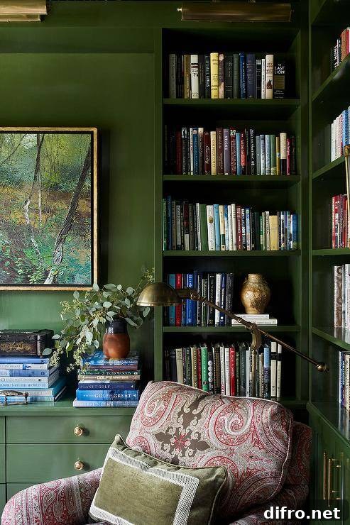 Welcoming home office features glossy green lacquered bookcases lit by brass picture lights and fitted with a brass swing arm sconce fixed above a pink and gray locked chair topped with a green velvet pillow. Art hangs from a green all over books stacked atop green drawers with brass pulls.
