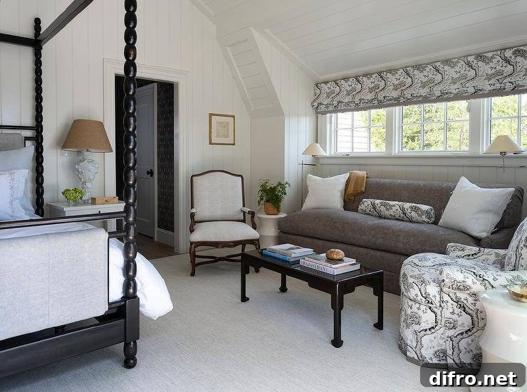 Cottagecore bedroom sitting area features a black chinoiserie coffee table placed on a gray rug in front of a gray damask couch topped with white pillows flanking a white and black pillows bolster pillow. The bolster pillow is made in fabric matching an upholstered accent chair and a white and black roman shade covering a row of windows flanked by polished nickel swing arm floor lamps. Walls and a vaulted ceiling are clad in shiplap trim.