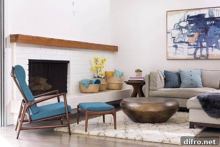 Wonderfully designed living room features a large blue abstract art piece hung above a gray shelter back sectional topped with blue pillows and placed facing a round brass coffee table positioned on a Moroccan trellis rug. A brown and blue mid-century modern chair is placed at an angle in front of a white brick mid-century modern fireplace finished with a stained wood mantel.