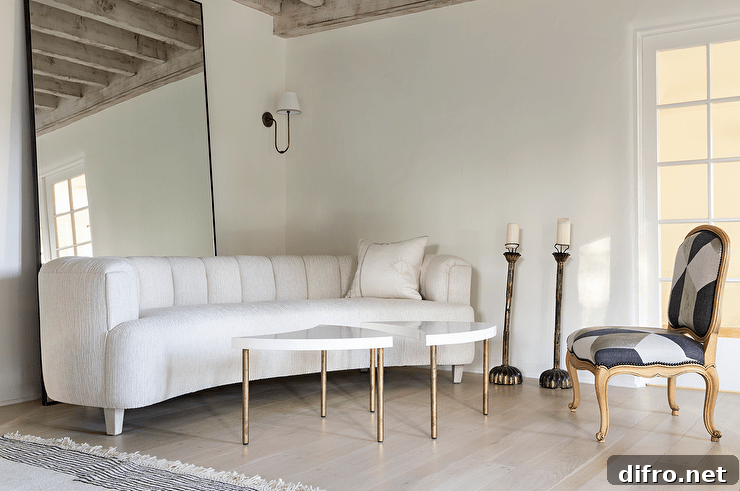 Living room features a white curved channel tufted sofa in front of a full length mirror, white and gold accent tables, a white, black and gray French chair and tall candle floor stands.