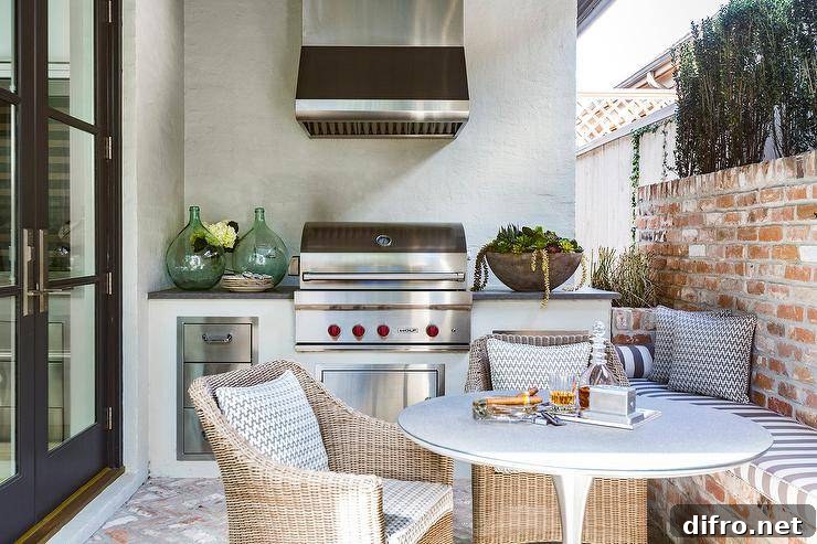 Outdoor patio designed with a red brick bench and wall upholstered with a gray and white striped cushion. A round outdoor dining table is paired with a set of wicker chairs next to a cooktop designed with a small wolf bbq, stainless steel vent hood, and kitchen space.