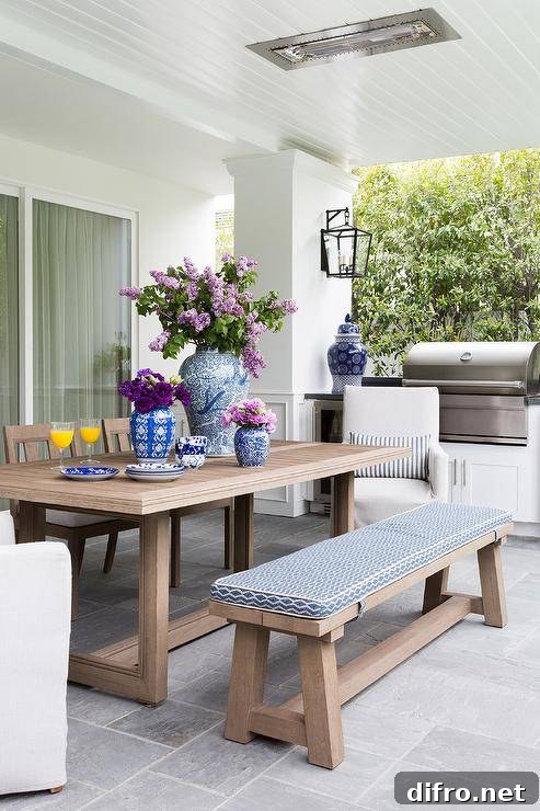 Brown teak dining table and a matching dining bench feature a blue and white pattern upholstered cushion. Natural linen slipcovered dining chairs mix with the bench and teak chairs atop slate pavers next to a covered patio bbq.