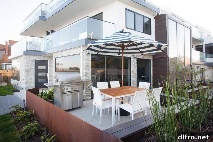 California beach style patio features a white dining table surrounded by white modern dining chairs shaded by a black and white striped umbrella while a stainless steel grill sits on a concrete floor.