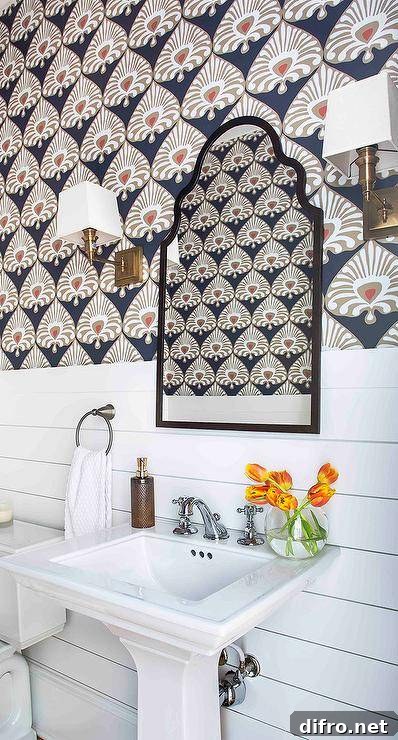 Between brass sconces, a black arch mirror is hung from a wall clad in taupe and blue wallpaper lined with shiplap trim. The mirror is mounted over a white pedestal sink finished with a polished nickel cross handle faucet kit.