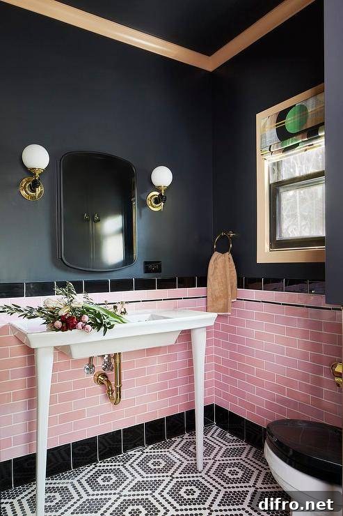 Gold and black sconces are mounted to a black painted wall on either side of a curved vanity mirror hung above pink offset wall tiles finished with black border tiles. A white French sink vanity is fixed in front of the backsplash tiles and against black and white hexagon floor tiles.