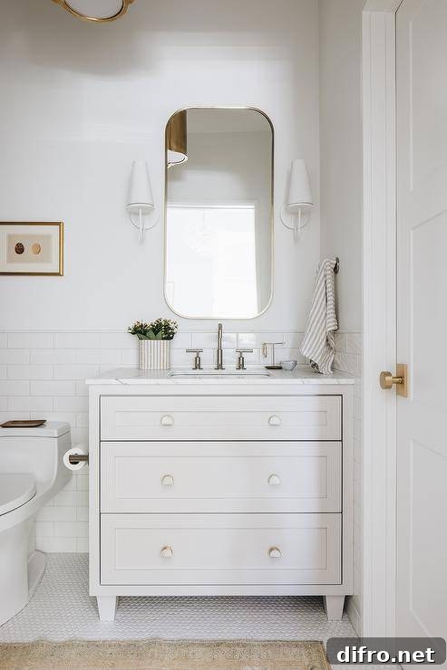 Aerin Fontaine Single Sconce flank a curved white mirror hung over an off-white dresser-like washstand finished with a polished nickel gooseneck faucet and white and gold half moon knobs. The washstand is mounted in front of a white subway backsplash tiles.