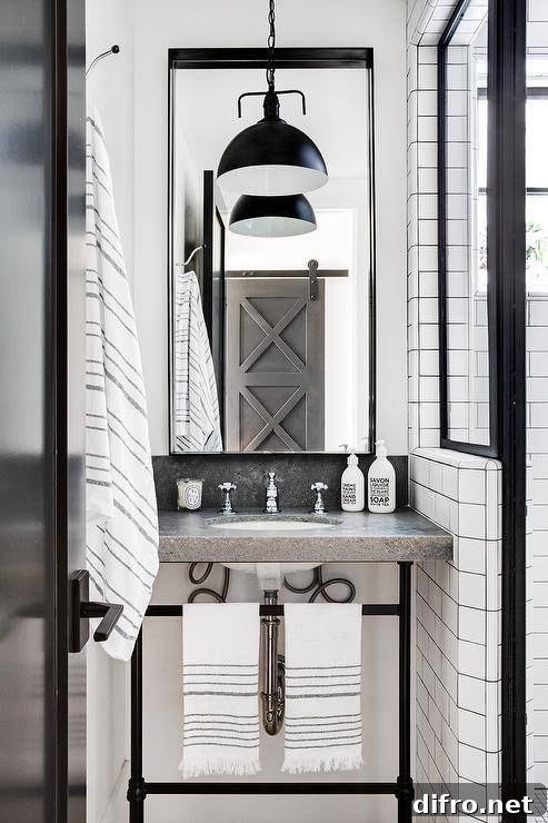 A concrete top washstand accented with oil rubbed bronze legs is fitted with a polished nickel cross handle faucet mounted beneath a tall black framed mirror illuminated by a black industrial pendant.