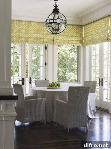 Sunshine filled dining room with dark wood floors, picture windows and French doors. The doors and windows are dressed in roman blinds with Kelly Wearstler Imperial Trellis Citrine fabric. The round skirted dining table pairs with four contemporary woven dining chairs. A sphere shaped glass and iron pendant hangs over the round table.