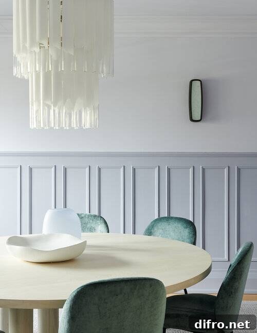 Half wainscot dining room walls are positioned behind a round cream dining table matched with green velvet dining chairs and lit by a white cascading chandelier.