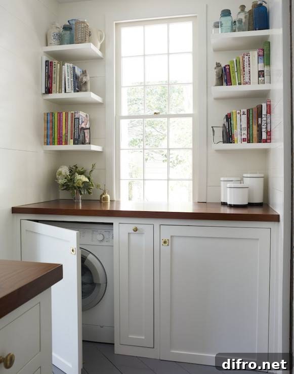Beautiful laundry room features small stacked floating bookshelves flanking double hung window over concealed washer and dryer hidden behind cabinet doors and topped with wood countertop.