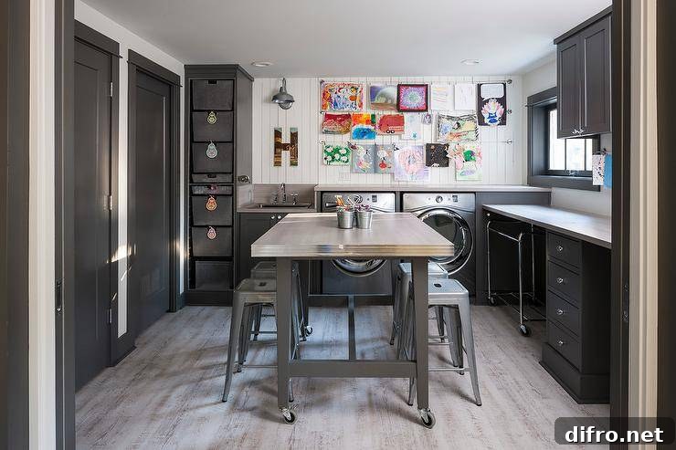 Tolix stools and a stainless steel rolling craft island centered in a gray laundry room doubling as a craft room. Black shaker cabinets frame the perimeter walls finished with white vertical planks displaying kids art on wires. Gray wash wood plank floors lighten the atmosphere of the room and creates a well balanced, decorative and practical space.