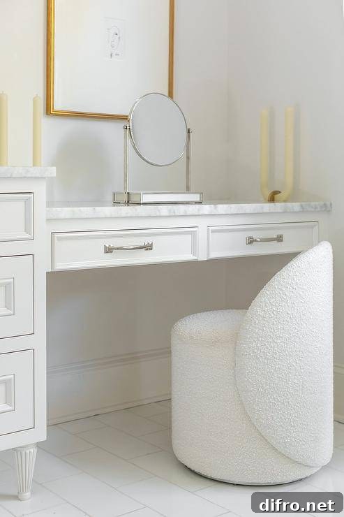 A white boucle chair sits on a white staggered floor tiles at a white drop-down makeup vanity finished with a marble countertop fixed under stacked art.
