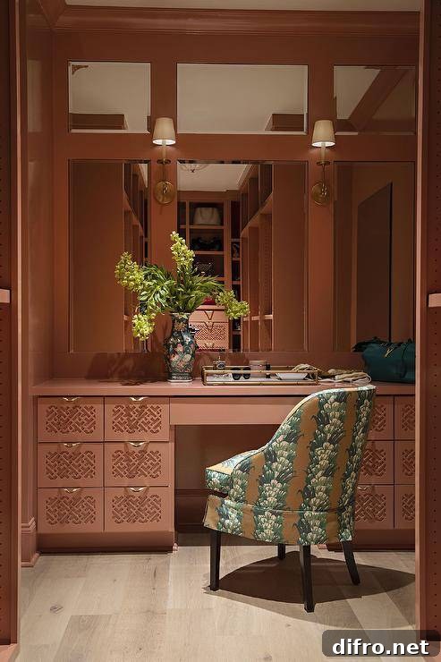 Paneled mirrors are lit by brass sconces fixed to a pink wall over a pink makeup vanity accented with lattice drawers fronts and brass pulls. The vanity is paired with a green and brown chair.