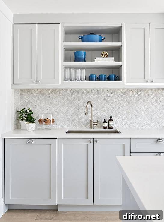 Small marble herringbone backsplash tiles add a decorative finish between light gray shaker cabinets with satin nickel cup pulls.