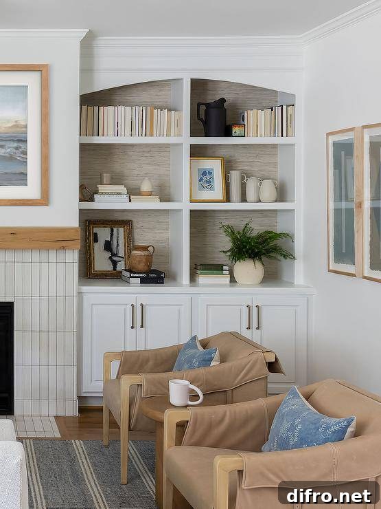 Brown leather flap accent chairs topped with blue pillows sit on a blue stripe jute rug on either side of a round wooden accent table. Gray wallpaper accents the back of an arched built-in bookcase mounted over white cabinets.