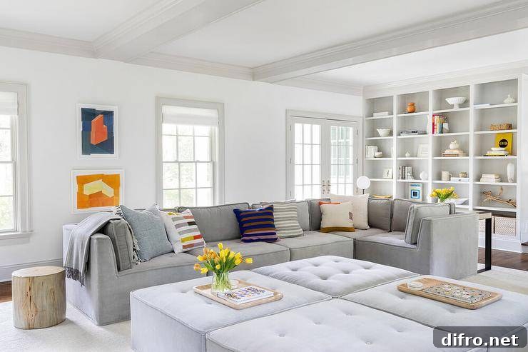 Gorgeous gray living room features a low gray tufted modular sectional placed beside a tree stump end table facing four gray tufted modular ottomans. Abstract blue and orange art hangs beside the sofa and between windows, while styled white built-in cabinets are fixed into white walls lined with light gray trim.