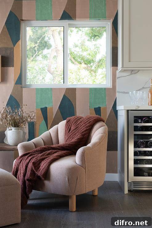 Earth toned geometric wallpaper frames a window located over a mauve tufted accent chair finished with wooden legs and draped with a brown throw blanket.