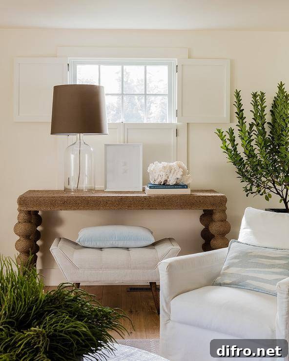 Anthropologie Jute Wrapped Console Table surely makes a statement in a cottage living room showcasing a glass lamp with a brown shade and a cream tufted bench underneath. Indoor plants white window trim and white surrounding furnishings create a dwelling sure to appeal the decorative eye and offer a place to rest and unwind.