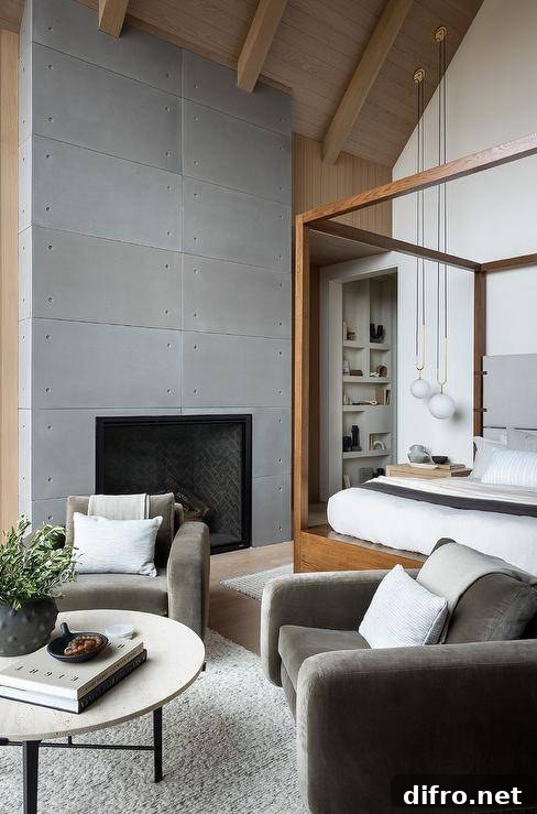 Bedroom features a sitting area with dark gray velvet accent chairs at at round black and white coffee table atop a gray rug, a modern fireplace and a wooden canopy bed under vaulted ceiling.