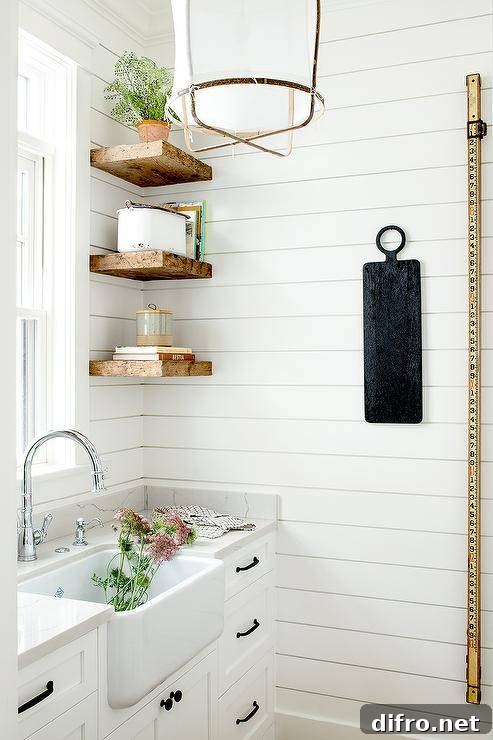 Stacked rustic wooden floating shelves are mounted against a shiplap backsplash over white kitchen drawers contrasted with oil rubbed bronze pulls. A farmhouse sink with a chrome gooseneck faucet is mounted beneath a window in a cottage-style pantry.