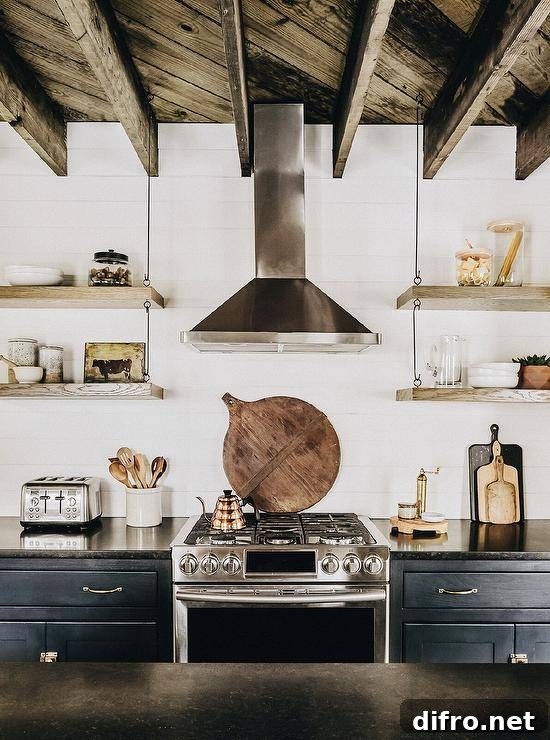 A rustic plank ceiling with rustic wood beams is mounted over a black and white kitchen featuring a stainless steel hood fixed to shiplap trim between wood and wire shelves. The shelves hang over black cabinets donning brass pulls and a black marble countertop flanking a stainless steel oven range.