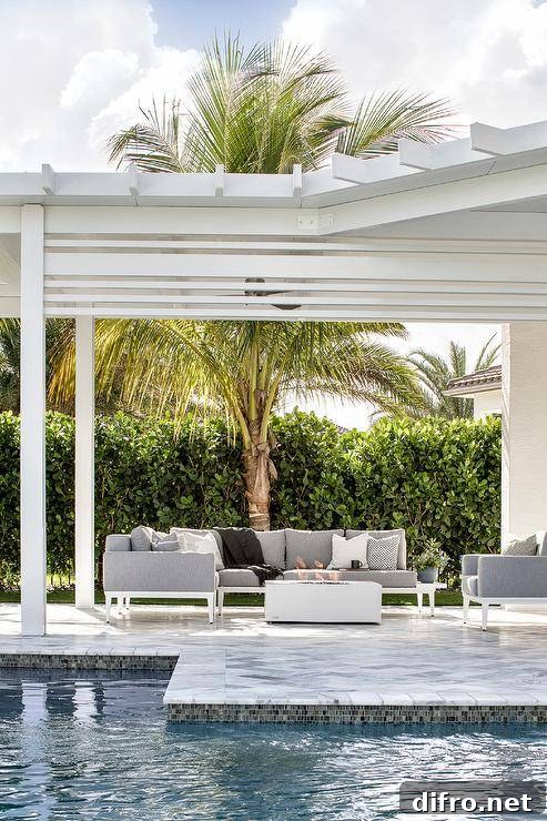 A white and gray outdoor sectional with white fire pit sits under a white pergola next to an in ground pool.