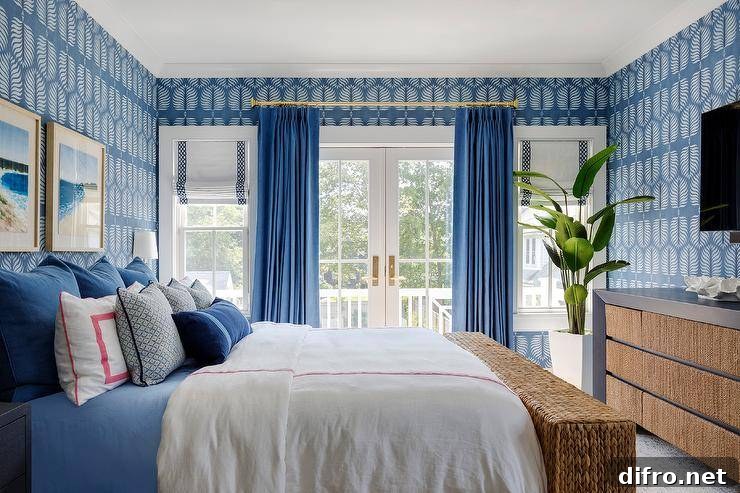 Restful blue bedroom features a bed dressed in red and blue bedding complemented with layered red and blue pillows. A seagrass bench sits at the foot of the bed and in front of a blue wooden dresser positioned under a TV hung from a wall covered in blue botanical wallpaper.