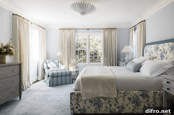 White and blue bedroom boasts a brass floor lamp and a white lattice stool placed beside a blue striped chaise lounge positioned on a blue rug in a corner between windows dressed in ivory curtains.