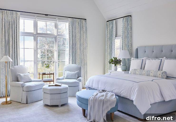 Blue primary bedroom design features a blue oval bench at the foot of a tufted bed with blue border shams, a white wooden nightstand lit by a blue lamp and a sitting area with light blue chairs.